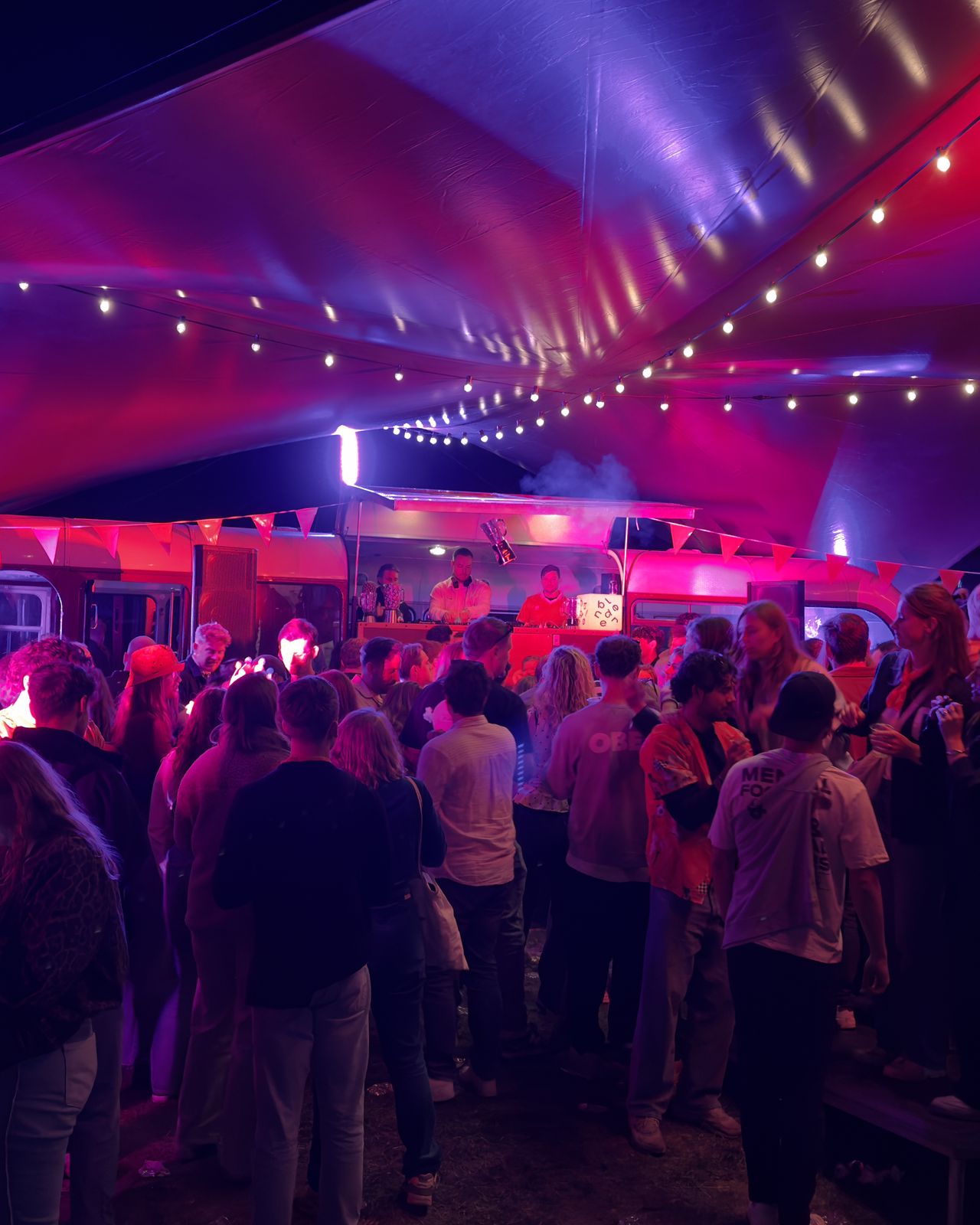 Volle tent in roze en rood licht met De Bus als DJ booth op Koningsdag Festival